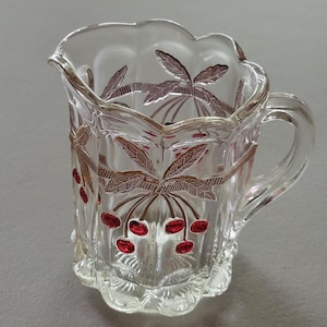 May include: A clear glass pitcher with a scalloped rim and a handle. The pitcher is decorated with a red and gold cherry design.
