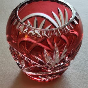 May include: A round, deep red cut-to-clear crystal bowl featuring intricate starburst and fan-shaped patterns etched into the glass. The rim has a scalloped edge, and the clear glass base contrasts with the vibrant red overlay, creating a faceted, reflective surface that catches the light.