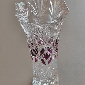 May include: A clear crystal vase with a flared top and a detailed, cut-glass design. The vase features a pattern of leaves and geometric shapes, with a band of purple accents near the middle. The vase is approximately 6 inches tall.