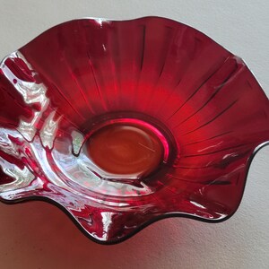 May include: A vibrant red glass bowl with a ruffled edge. The bowl has a circular base and a flared, petal-like rim. The glass is translucent, allowing light to pass through, and has a glossy finish.