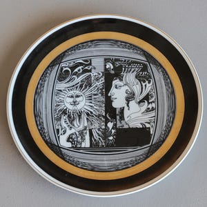 May include: A black and white ceramic plate with a gold rim. The plate features a stylized design of a sun and a woman's face.