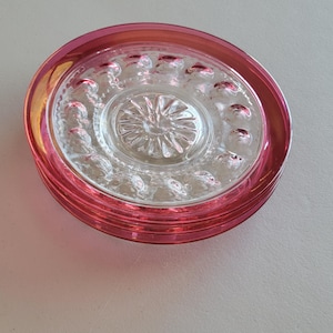 May include: A stack of clear glass coasters with a pink rim. The center of each coaster has a starburst design. The rim has a series of small, raised dots. The coasters are circular and appear to be stacked on top of each other.