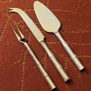 May include: A silver cheese fork, knife, and cake server set with bamboo-style handles. The set is displayed on a textured, reddish-brown surface. The utensils have a shiny, reflective finish.