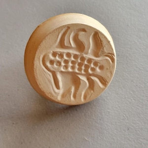 May include: A round, light brown clay stamp with a detailed scorpion design. The stamp features a handle and a raised relief of the scorpion's body, claws, and tail. The scorpion's body is segmented with small, circular details.