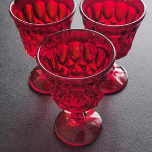 May include: Three ruby red, stemmed glasses with a diamond-cut pattern. The glasses have a rounded bowl and a short stem, perfect for serving a small beverage. The glasses are arranged on a dark surface.