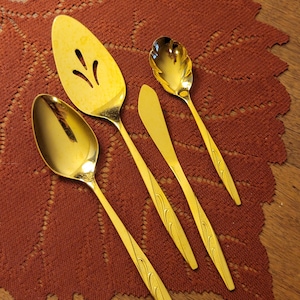 May include: A set of four gold-colored serving utensils, including a slotted pie server, a serving spoon, a butter knife, and a ladle. The utensils are arranged on a rust-colored, leaf-shaped placemat, set on a wooden surface.