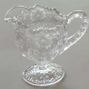 May include: A clear glass creamer with a footed base and a floral design. The creamer has a handle and a scalloped rim.