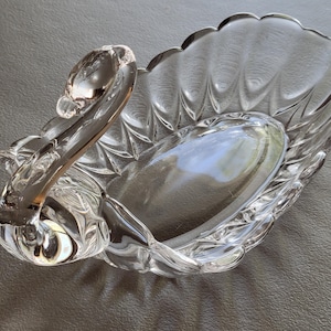 May include: Clear glass swan-shaped dish. The dish features a swan's head and neck as a handle, with a scalloped edge and a textured, oval-shaped bowl. The glass is transparent, reflecting light and creating a shimmering effect.