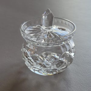 May include: A clear, cut-glass jar with a lid. The jar has a round body with a diamond pattern and a pointed finial on the lid. The jar is designed to hold small items and is made of crystal.