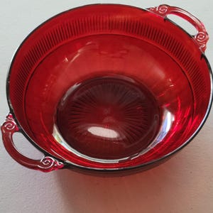 May include: A red glass bowl with two handles. The bowl has a ribbed design and is made of ruby red glass.