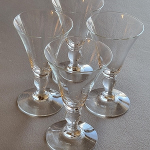 May include: Four clear glass cordial glasses. Each glass has a round base, a decorative stem, and a flared bowl. The glasses are arranged in a slightly overlapping pattern, showcasing their elegant design and clarity.