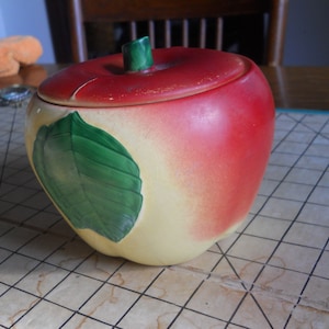 May include: A ceramic apple-shaped jar with a lid. The jar is primarily red with a yellow base and a green leaf design. The lid has a green stem. A decorative kitchen item.