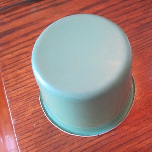 May include: A light green enamelware pot with a rounded body and a white rim. The pot is sitting on a wooden surface. The pot is a classic kitchenware item.