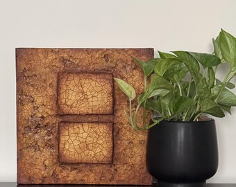 May include: A square, brown, textured wall art piece with two rectangular sections. The sections have a cracked, aged look. The art piece is on a dark brown surface next to a potted plant with green leaves.