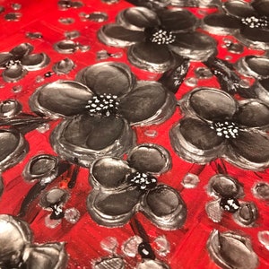 May include: Close-up of a red canvas painting with black and silver flowers. The flowers are painted in a textured style, giving them a 3D effect.