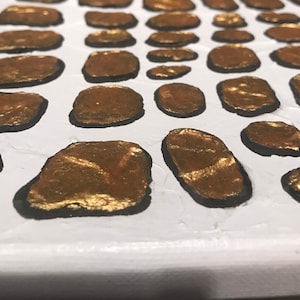 May include: Close-up of a white canvas with a pattern of gold and black oval shapes. The gold shapes are painted with a textured, metallic finish.