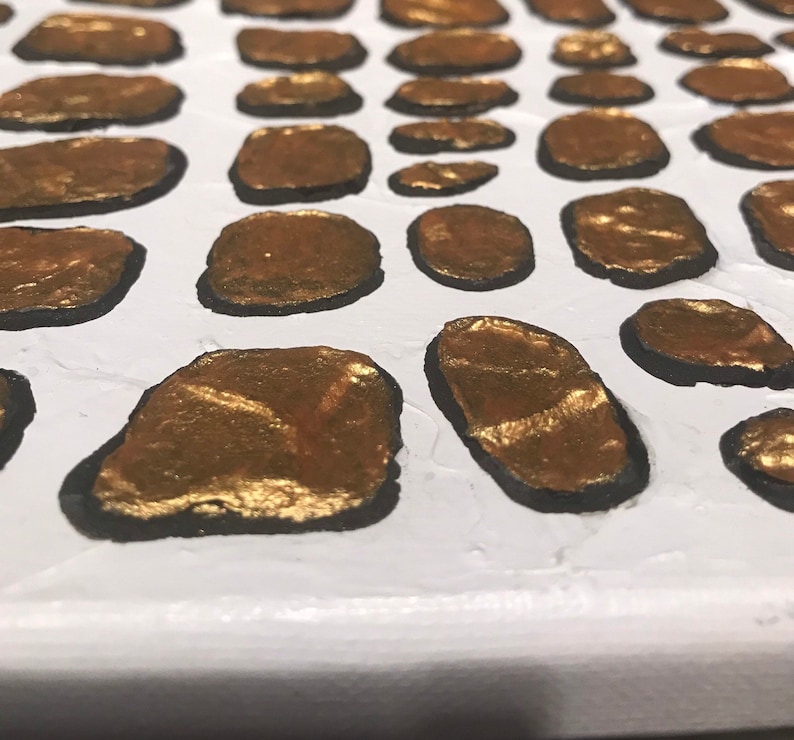 May include: Close-up of a white canvas with a pattern of gold and black oval shapes. The gold shapes are painted with a textured, metallic finish.
