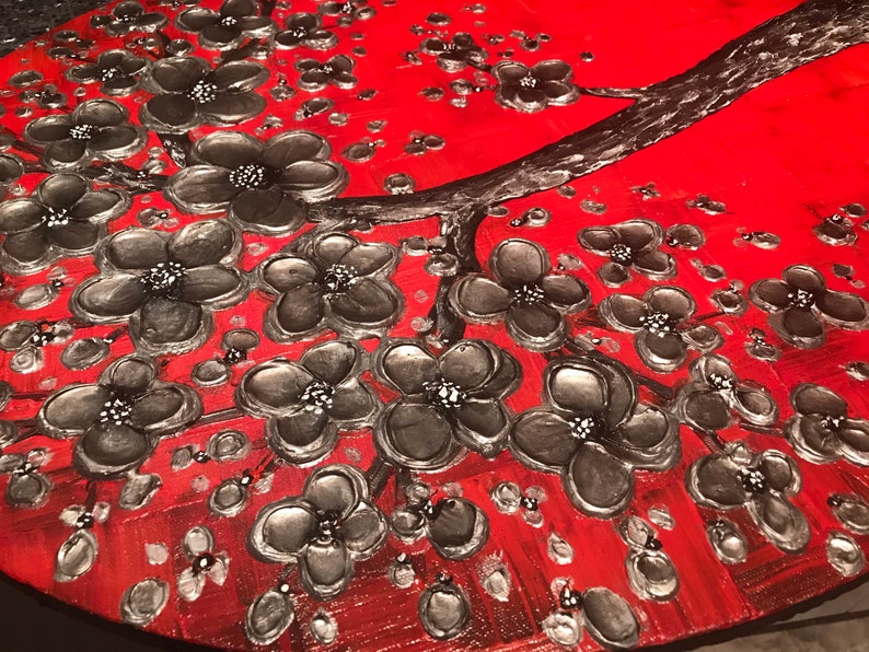 May include: A close-up of a red and black abstract painting featuring a stylized tree with silver-colored flowers. The flowers are painted in a textured style, giving the painting a three-dimensional effect.