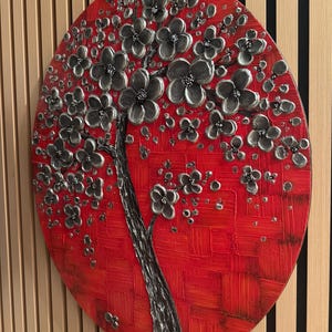 May include: A round canvas painting with a textured red background and a silver 3D tree with black flowers. The tree is in the center of the canvas and the flowers are blooming.