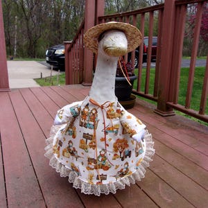 May include: A white goose statue wearing a straw hat and a fall-themed dress. The dress features a print of scarecrows, pumpkins, and autumn foliage. The dress is trimmed with a white lace.