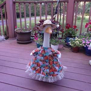 May include: A decorative goose statue wearing a straw hat and a colorful dress with a pumpkin pattern. The dress has a white lace trim. The goose is standing on a wooden deck with potted plants in the background.