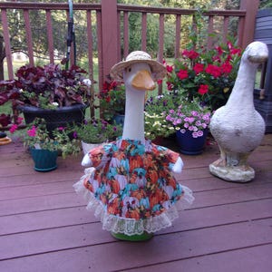 May include: A white goose statue wearing a straw hat and a dress featuring a colorful pumpkin print with a cream-colored lace trim at the hem. The goose has an orange beak and is positioned upright, showcasing the detailed fabric pattern.