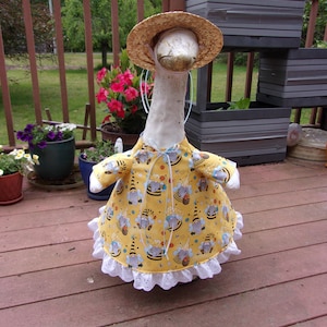 May include: A decorative goose figurine wearing a yellow dress with a bee and gnome pattern, a straw hat, and a white lace trim. The goose is standing on a wooden deck, with potted plants in the background.