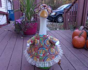 May include: A decorative goose statue wearing a brown dress with a colorful paisley pattern, a matching hat, and a white lace trim. The goose has a long white neck and a yellow beak. Two orange pumpkins are in the background.