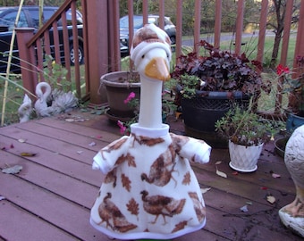 May include: A white goose figurine wearing a brown and white hat and a matching dress with a turkey and leaf pattern. The goose is standing on a wooden deck, surrounded by potted plants.