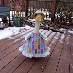 May include: A goose figurine wearing a straw hat and a colorful dress with a bunny pattern. The dress has a white lace trim. The goose is standing on a wooden deck.