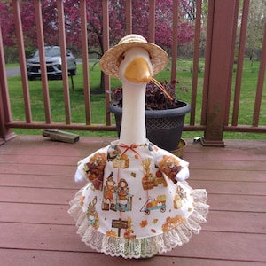 May include: A white goose wearing a straw hat and a fall-themed dress. The dress features a print of pumpkins, scarecrows, and autumn leaves. The dress has a lace trim.