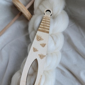 May include: A wooden weaving tool with a handle and an oval opening, featuring engraved bee designs. The tool has text that reads "Eye, Fine, Spun, Ok, Worsted, Bulky, Super Bulky". The tool is resting on a bundle of white yarn.