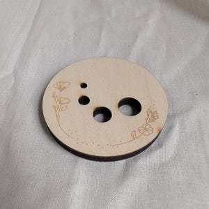 May include: A round, light-colored wooden coaster with three holes and floral etched designs. The coaster has a dark brown edge and is likely made of plywood. The floral designs are on the top surface of the coaster.