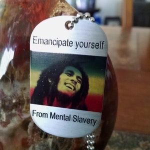 May include: Silver dog tag pendant with the text "Emancipate yourself From Mental Slavery" and a colour image of a smiling man. The pendant hangs from a silver ball chain. The background is blurred.