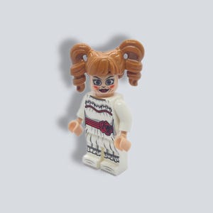 May include: A Lego figurine of a doll in a white dress with a red rose detail. The doll has light brown pigtails and a painted face with blue eyes and red lips. The figurine is standing on a white surface.