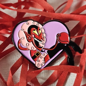 May include: A heart-shaped enamel pin featuring a red devil character with curly pink hair, yellow eyes, and a wide grin. The character wears a red outfit with a black belt and gold buckle, set against a light purple background within the heart frame.
