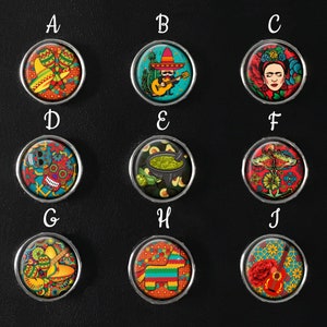 May include: Ten circular buttons with silver edges, each featuring a different vibrant design inspired by Mexican culture. Designs include a mariachi, Frida Kahlo, a sugar skull, guacamole, a piñata, and musical instruments. The background is black.
