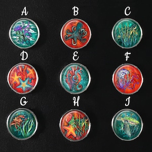 May include: A collection of ten round, silver-rimmed buttons with colorful underwater designs. Each button features a different marine creature or element, including jellyfish, octopus, seahorse, starfish, and a sea turtle. The designs are set against vibrant backgrounds.