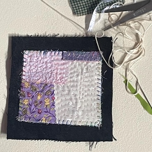 May include: A square fabric patch featuring a black border surrounding a patchwork center of white, light pink, and purple floral fabric. The pieces are joined with visible white stitching. Nearby are red-handled scissors, white thread, and pins.