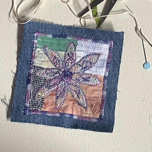 May include: A square denim fabric patch featuring a central embroidered purple flower motif on a multi-colored patchwork background. Red-handled scissors, loose white thread, sewing needles, and a small green and black checkered fabric scrap.