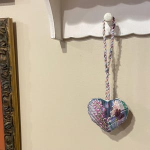 May include: A heart-shaped fabric ornament with a braided cord for hanging. The ornament features a patchwork design with floral embroidery and plaid fabric in shades of pink, purple, and blue. It hangs from a white shelf.