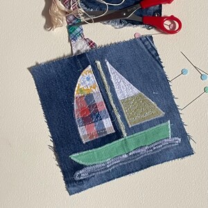 Peut inclure: Un carré de tissu en jean avec une application de voilier. Le bateau possède une coque vert clair, une voile blanche et vert olive, et une voile à carreaux multicolores. Des ciseaux à poignées rouges et des épingles sont posés à côté.