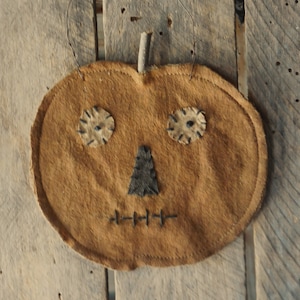May include: A flat, round, tan fabric pumpkin ornament featuring two circular patches for eyes, a triangular patch for a nose, and a horizontal stitched line for a mouth, all in dark thread. A small wooden twig serves as the stem with a wire hanger.