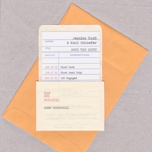 A library borrower’s card used as a wedding save the date announcement is resting in a customized library sleeve, and sits atop a Kraft catalog envelope. The background is gray.