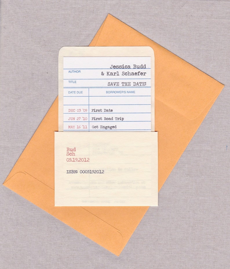 A library borrower’s card used as a wedding save the date announcement is resting in a customized library sleeve, and sits atop a Kraft catalog envelope. The background is gray.