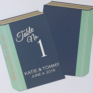 Puede incluir: Una tarjeta de n&uacute;mero de mesa con forma de libro azul y verde con el texto "Table No. 1" y los nombres "KATIE & TOMMY" y la fecha "JUNE 4, 2016". El lomo del libro tiene el texto "Writing Our Story" en dorado.