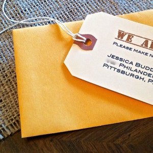 May include: A white luggage tag with brown text that reads "WE ARE MOVING PLEASE MAKE NOTE OF OUR NEW ADDRESS JESSICA BUDD & KARL SCOTT PHILANDER STREET PITTSBURGH, PA". The tag is tied to a yellow envelope with a brown string.