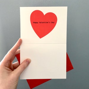 以下が含まれることがあります： 赤いハートと「Happy Valentine's Day」の文字が書かれた白いカード。カードは半分に折り畳まれており、赤い封筒が付いています。