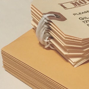 May include: A stack of brown envelopes with a stack of cream-colored tags tied together with white string. The tags have the text "I Must" and "Please" printed on them.