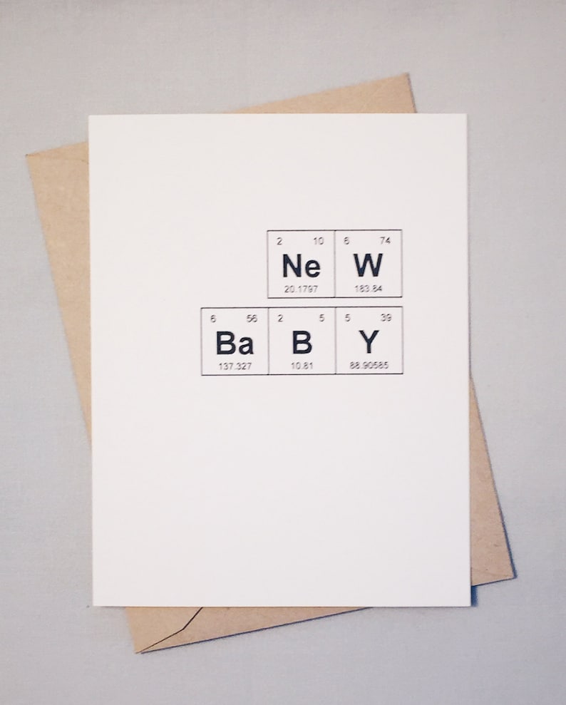 A white greeting card with the phrase “New Baby” spelled out using the Periodic Table of the elements. A Kraft envelope lays behind the card. The background is gray.
